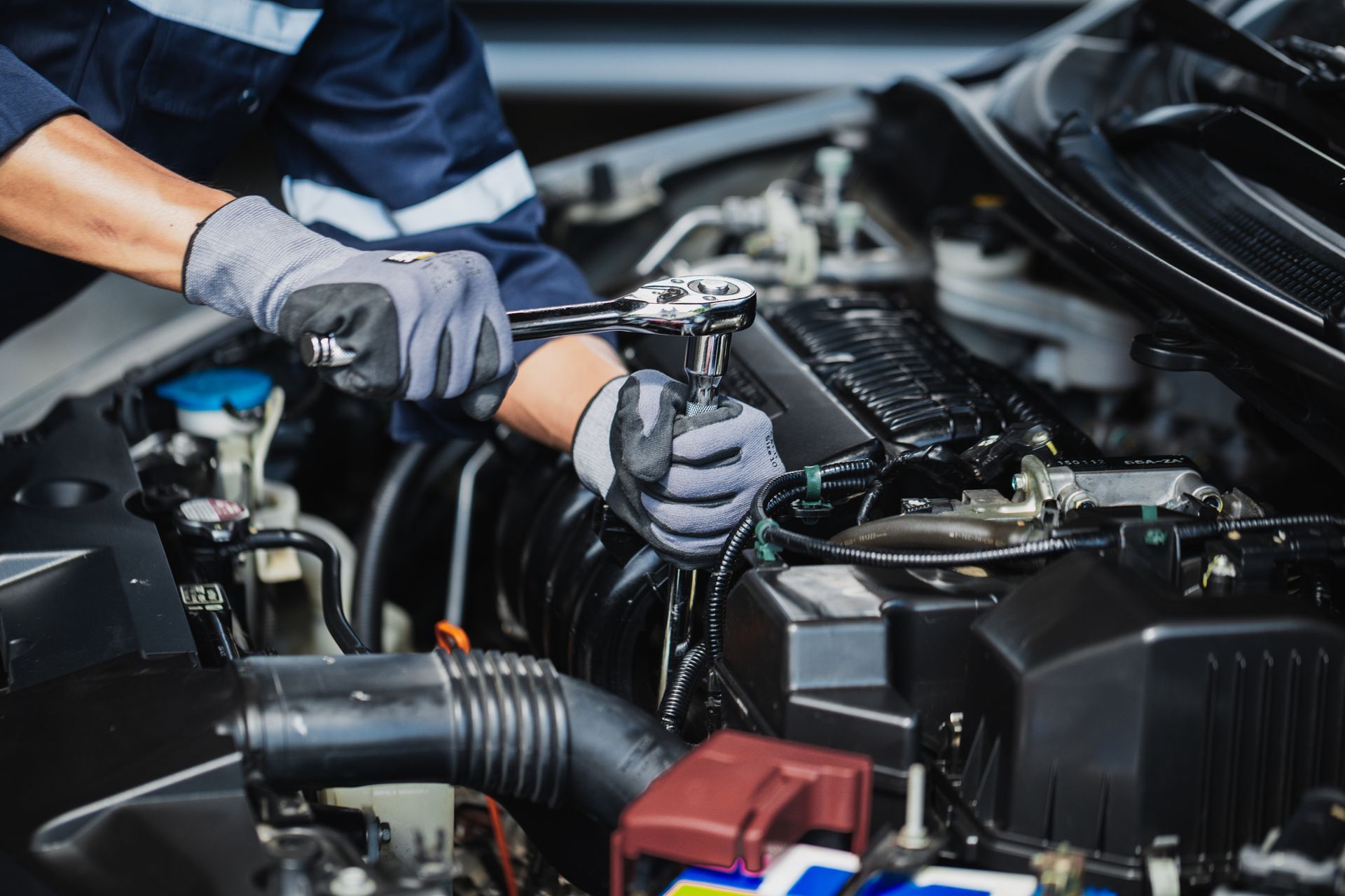 Mechanic using a wrench on a car engine, wearing gloves and a blue uniform.