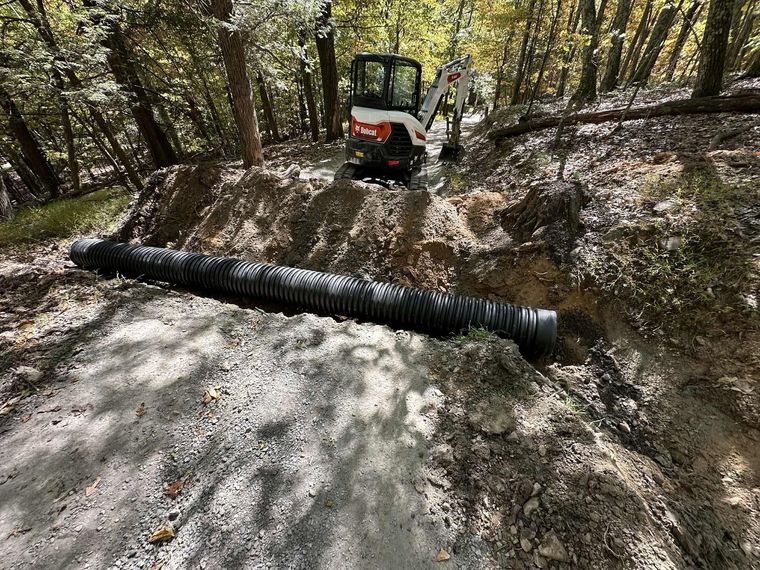 Small excavator on a muddy forest trail beside a large black drainage pipe