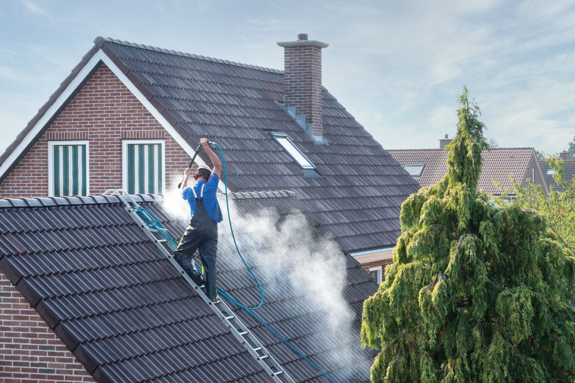 Person on a ladder power washing a dark gray shingled roof, creating steam; brick house, blue sky.
