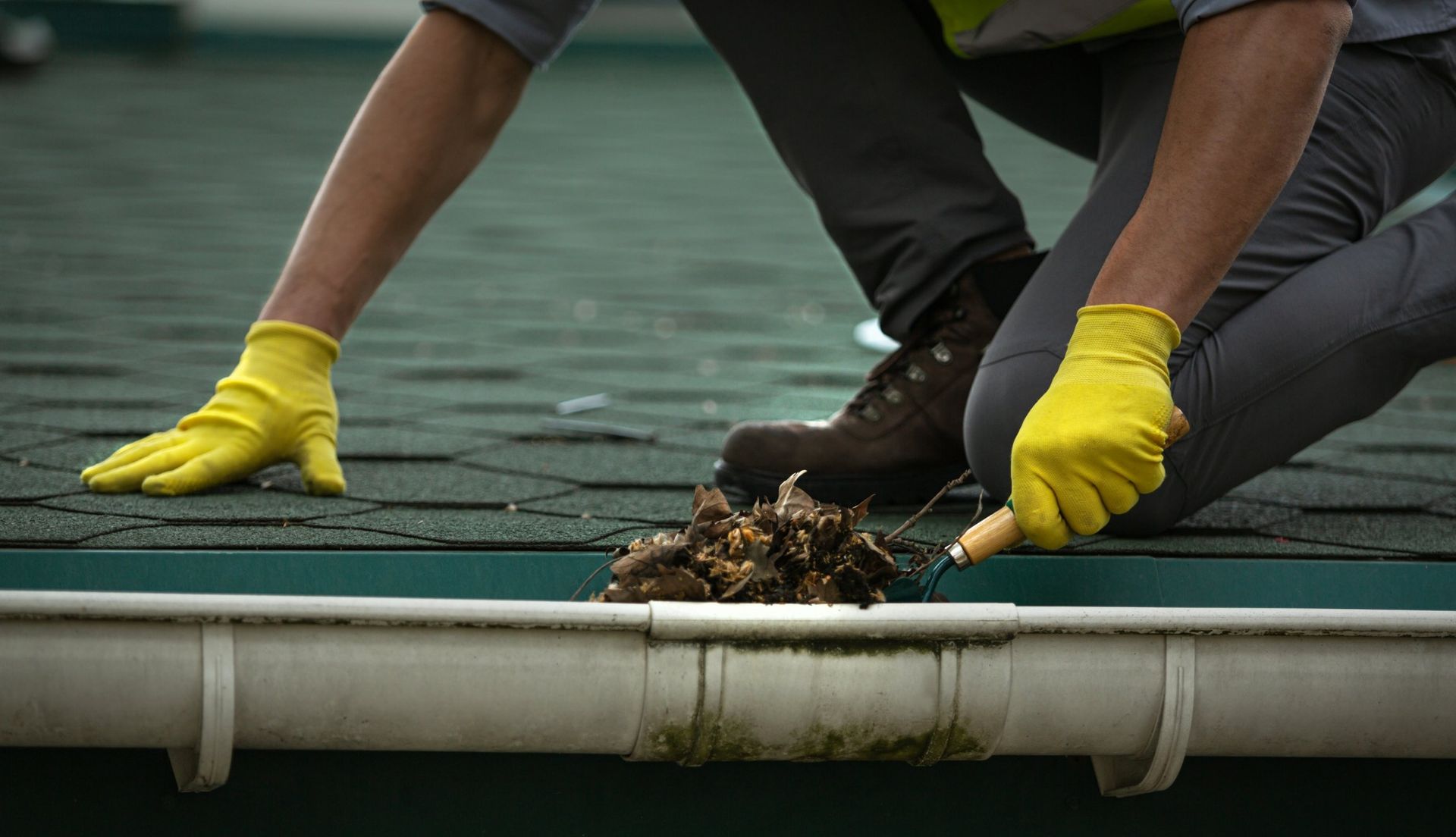 Person cleaning a gutter on a roof, wearing gloves, kneeled down, and holding a tool to remove debris.