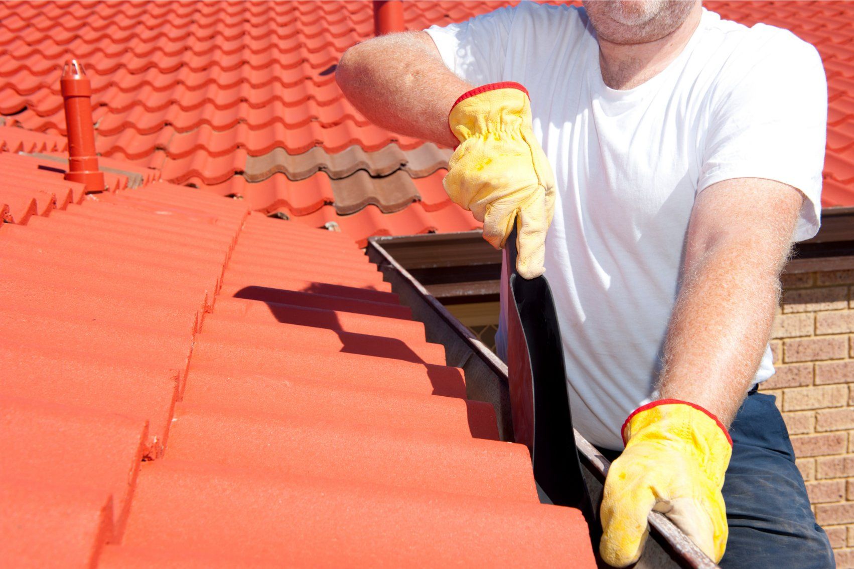 Man in yellow gloves cleaning a rain gutter on a red tiled roof.