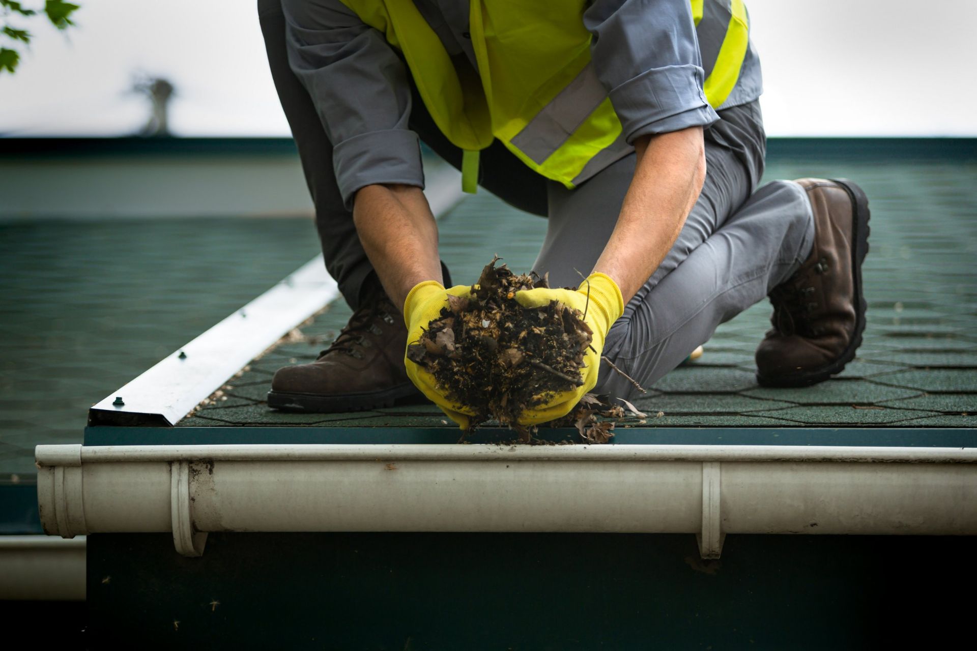 Person cleaning a gutter, kneeling on a roof with a yellow safety vest, holding debris.