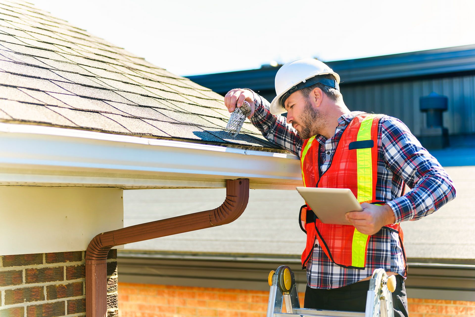 Man in hard hat and safety vest inspecting roof gutter with a tablet.
