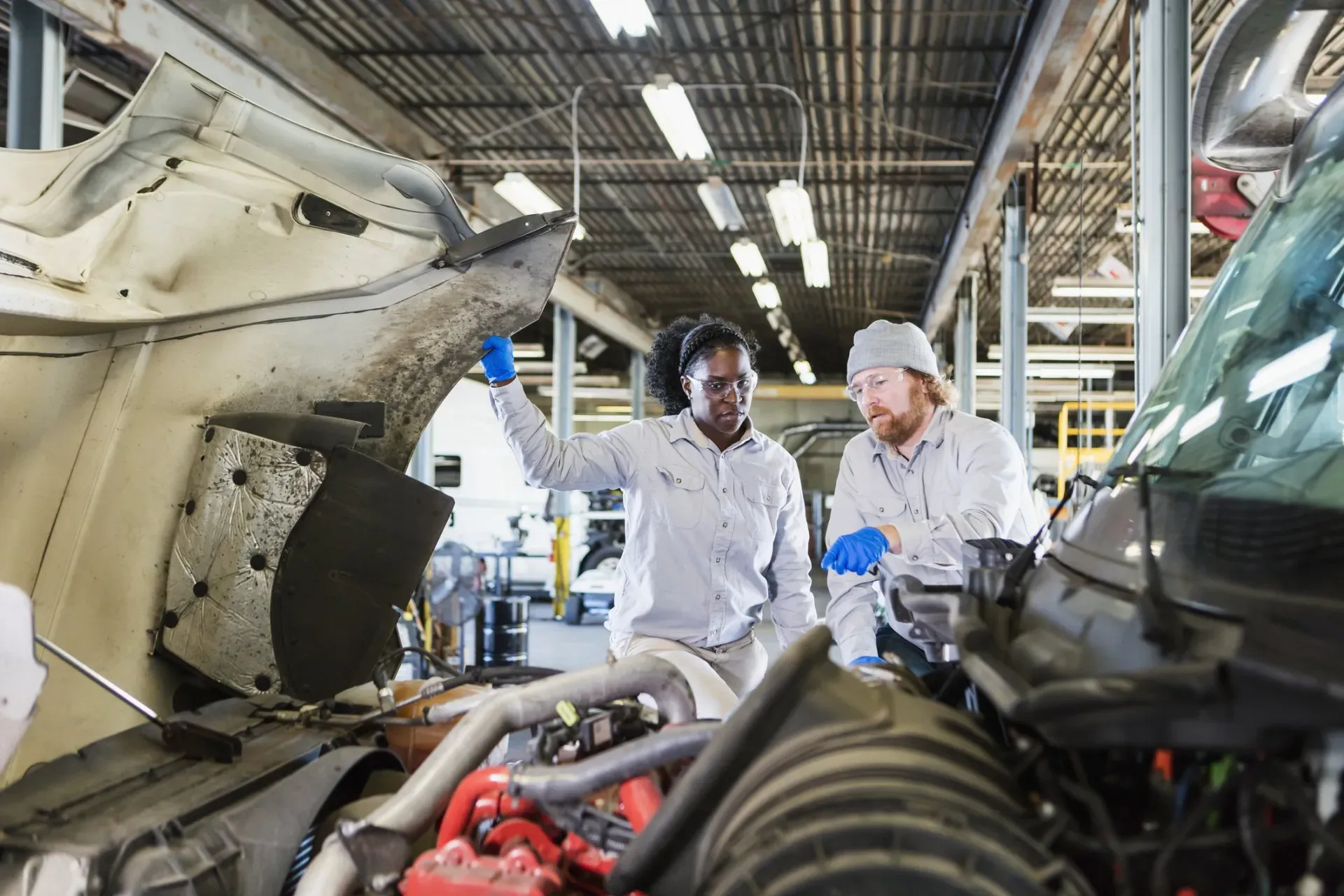 Two technicians wearing light-colored uniforms and blue gloves inspect the open engine bay of a heavy truck in a workshop. | Kensington Motor Cars