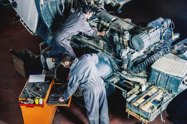 Two technicians in gray jumpsuits work on a large exposed vehicle engine inside a workshop. | Kensington Motor Cars
