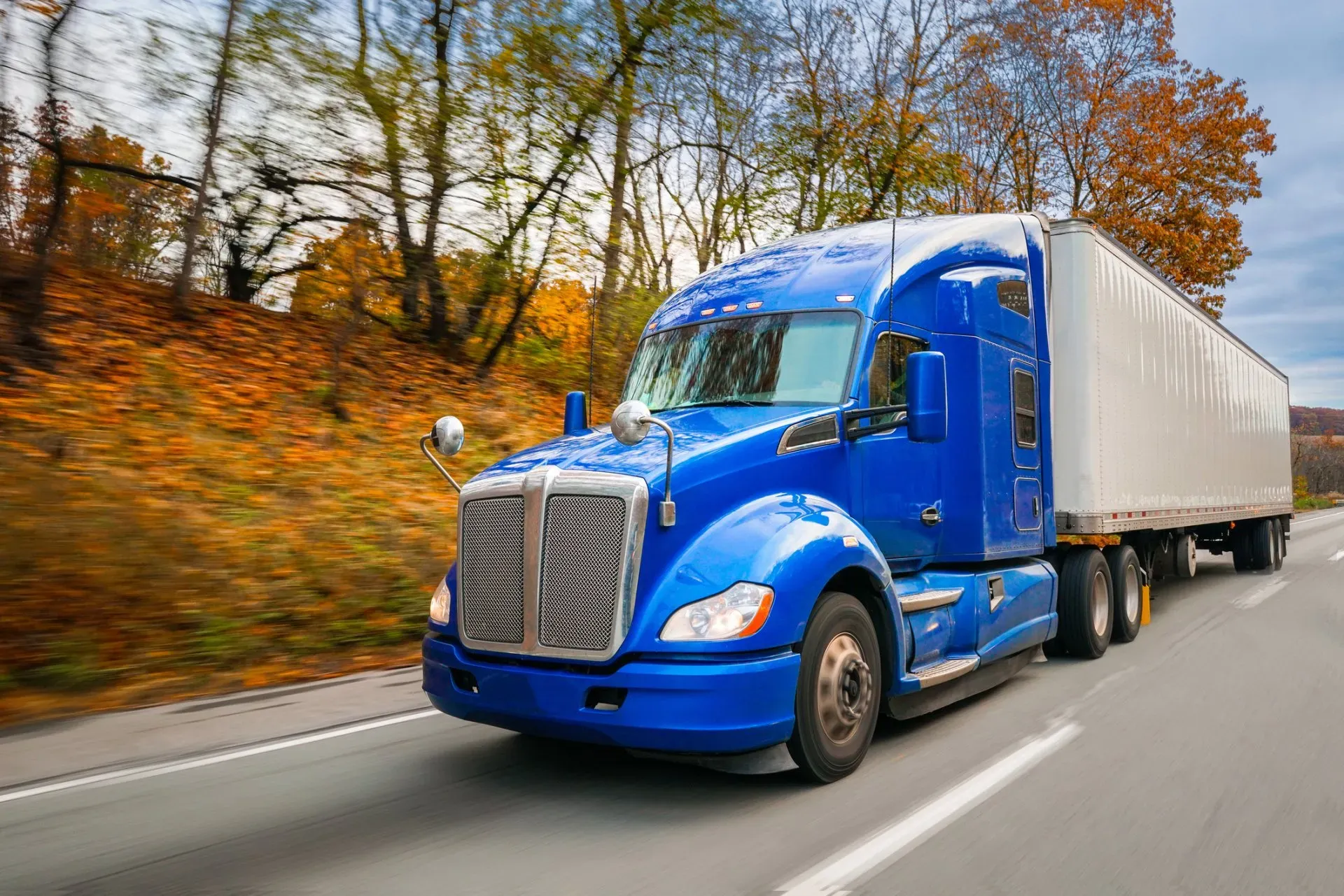 A bright blue semi-truck with a white trailer driving on a highway lined with autumn trees. | Kensington Motor Cars