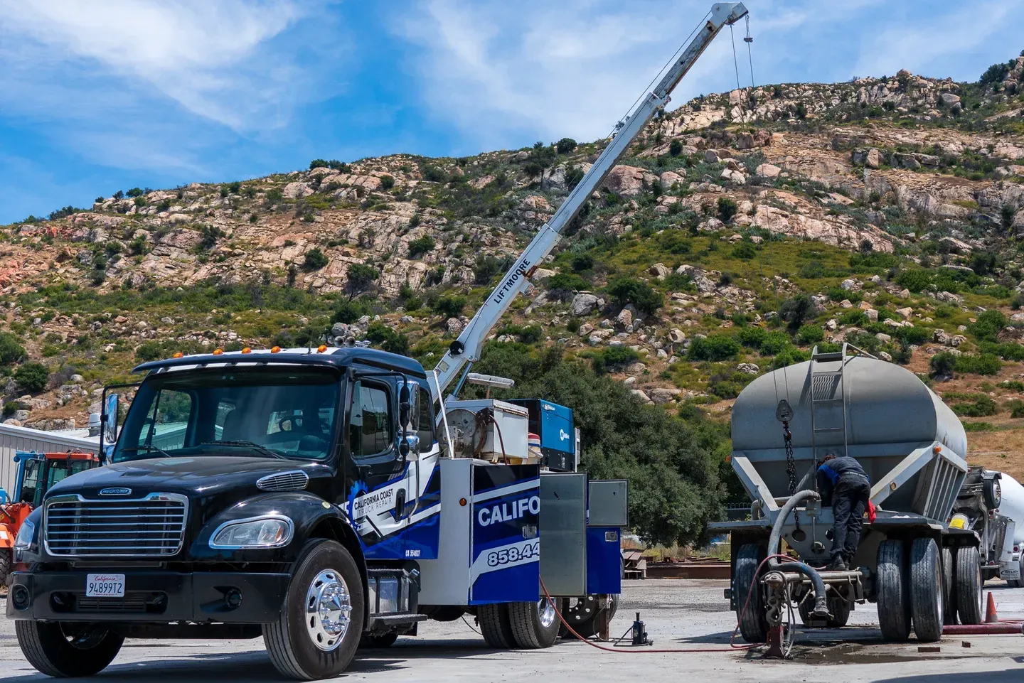 A black and blue service truck with a crane parked in a rocky, outdoor area next to a large industrial tank trailer. | Kensington Motor Cars