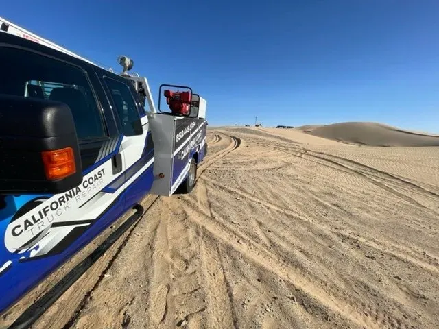A blue and white California Coast truck parked on a sandy desert dune under a clear blue sky. | Kensington Motor Cars