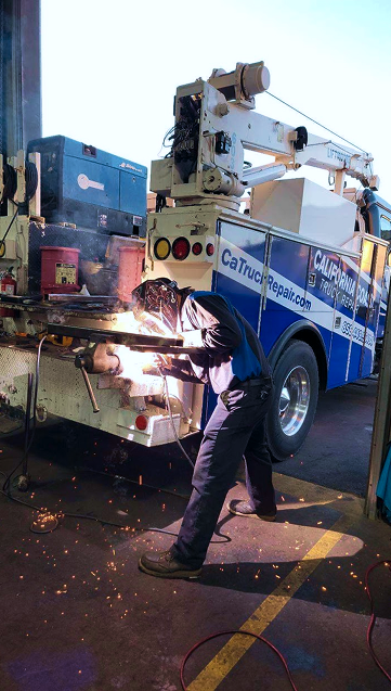 Person welding on a truck bed, sparks flying. Blue and white truck, crane in the background, outdoor setting. | California Coast Truck Repair