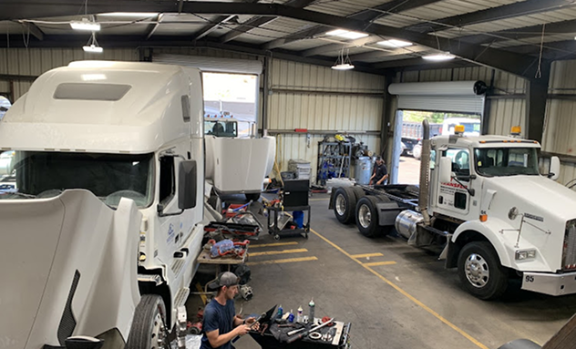 A person works on a semi-truck in an indoor repair shop where another truck is parked nearby. | California Coast Truck Repair