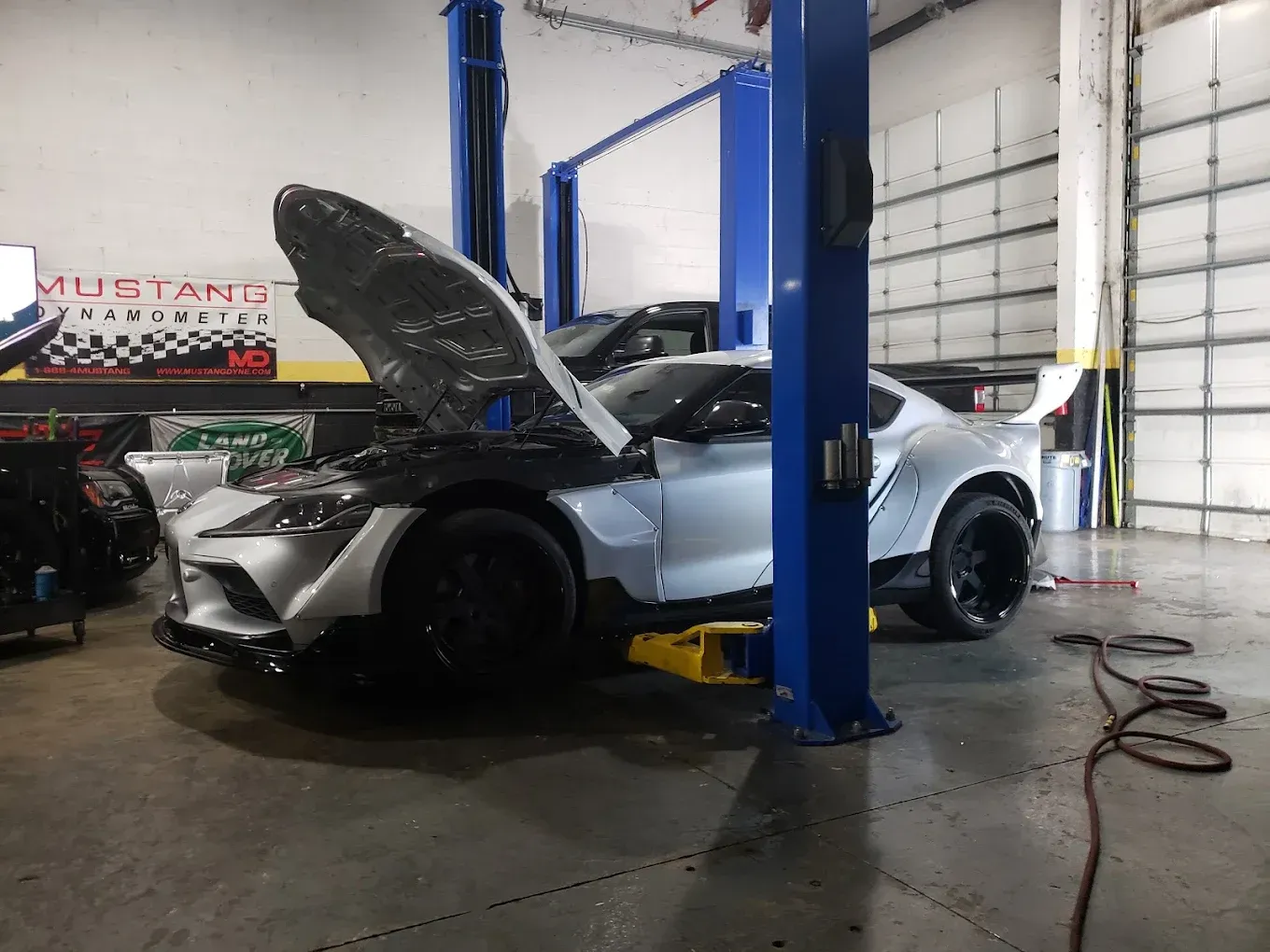 Silver sports car with open hood on a lift inside a mechanic shop. Blue lift posts and a roll-up door are visible.