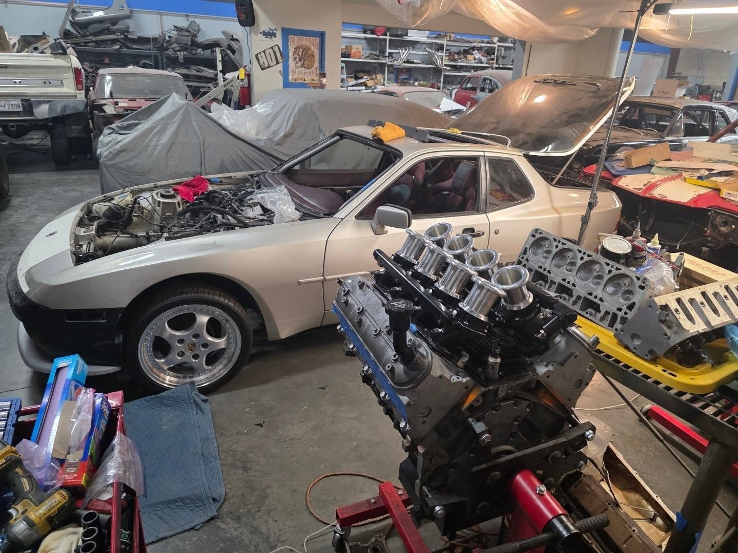 A white Porsche with hood open, near a disassembled engine in a garage.