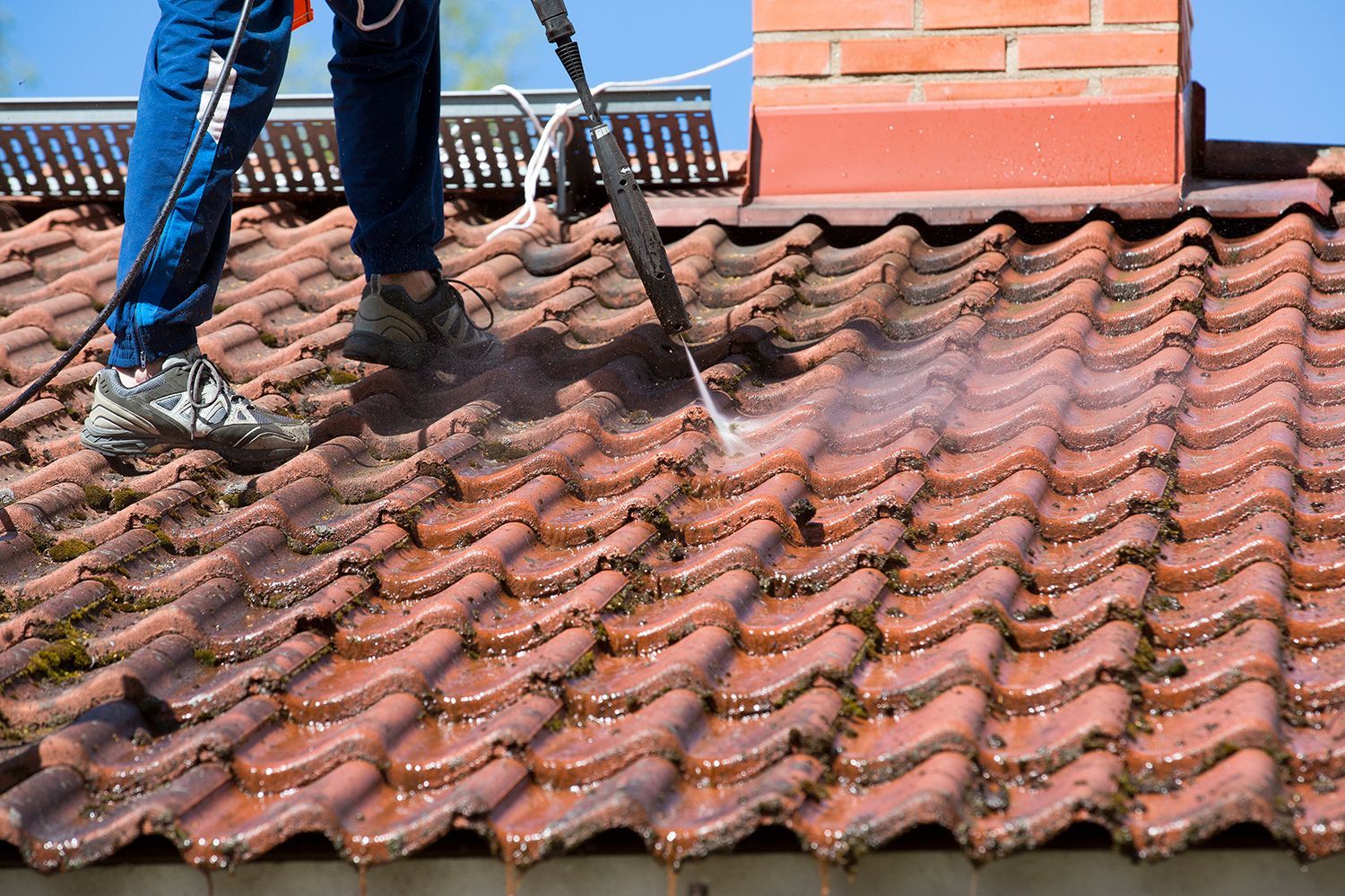 Worker cleaning moss from a terracotta-tiled roof with a brush near a brick chimney Worker cleaning moss from a terracotta-tiled roof with a brush near a brick chimney