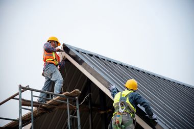 Two construction workers in hard hats installing roofing panels on a steep roof