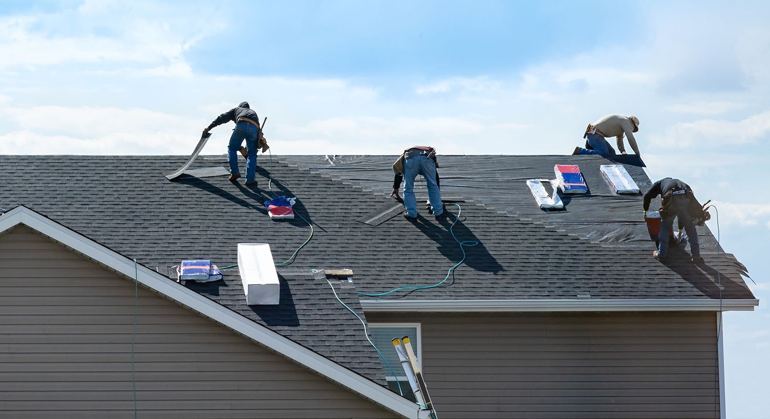 Workers repairing a shingled roof on a house, with tools and materials spread across the roof.