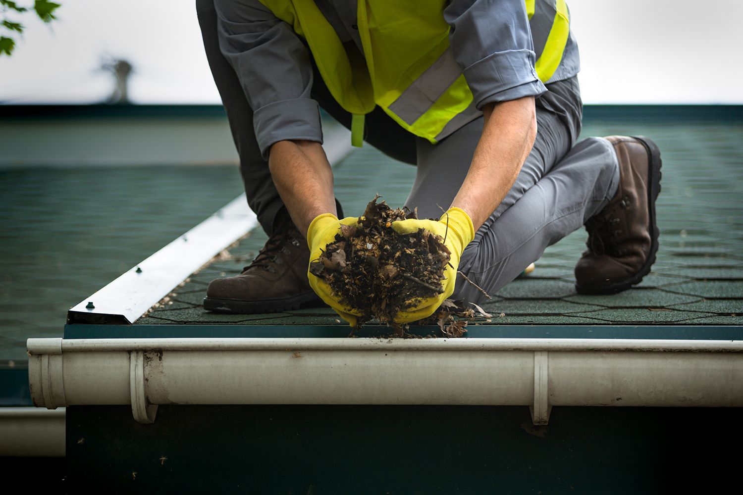 Worker in safety gear kneeling on a roof, cleaning or repairing a green shingle surface. Worker in safety gear kneeling on a roof, cleaning or repairing a green shingle surface.