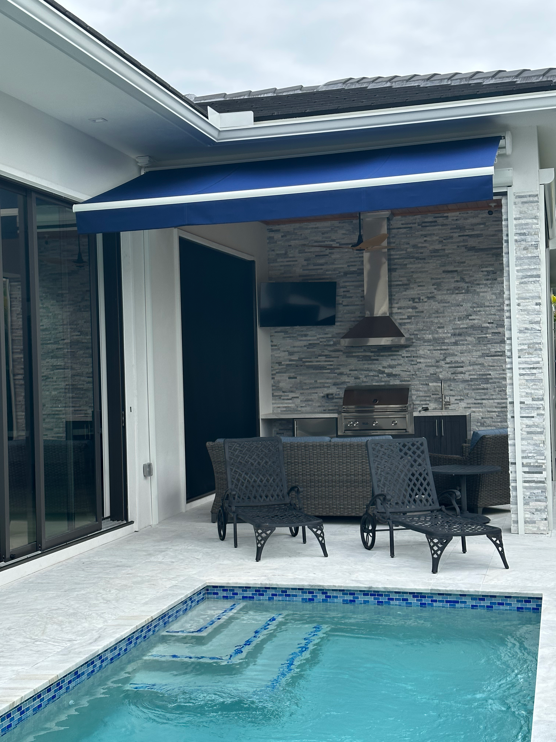 Poolside patio with blue awning, outdoor kitchen, seating, and pool with blue tile.