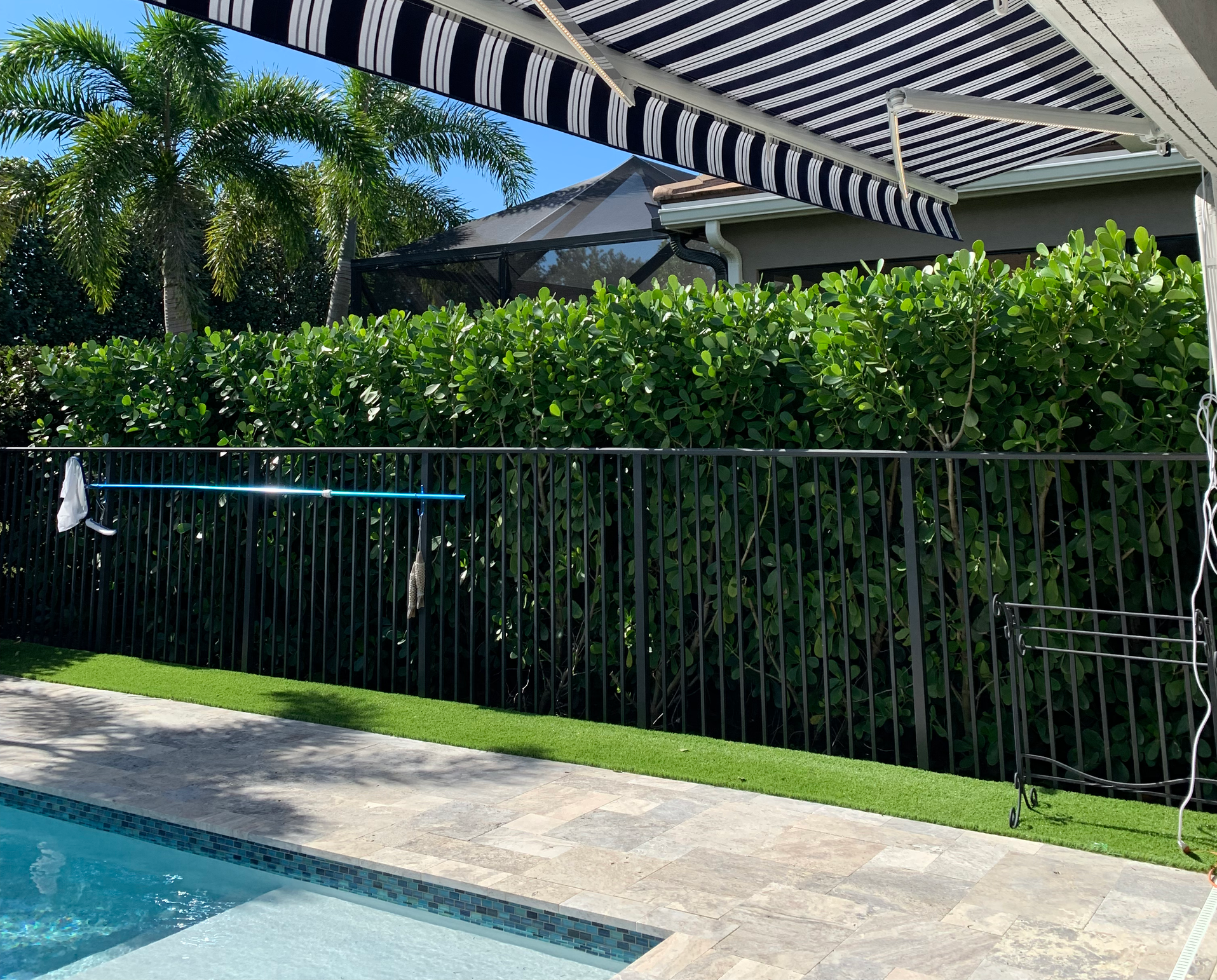 Poolside view with a black fence, lush green hedge, striped awning, and blue sky.