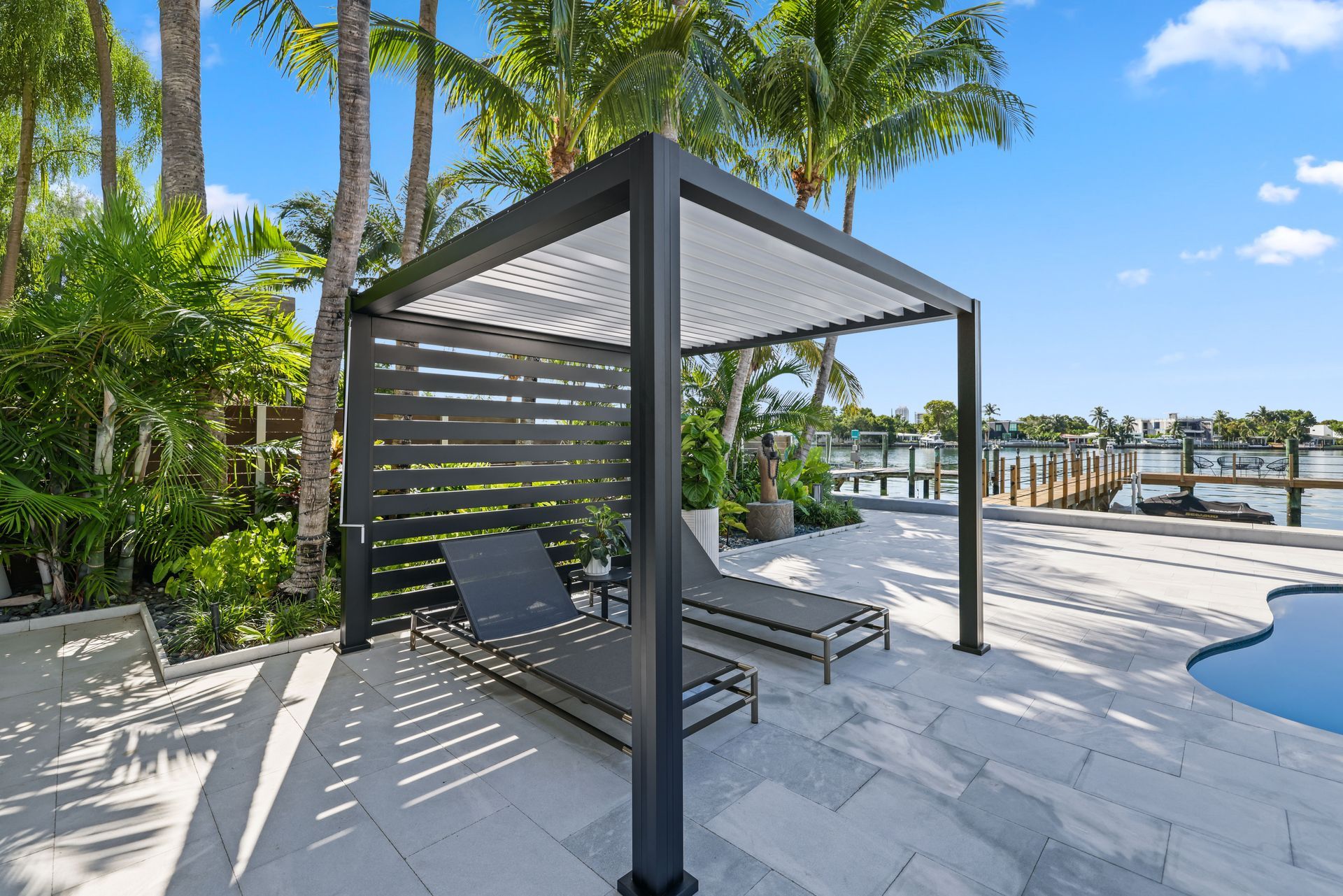 Lounge chairs under a black pergola on a stone patio, overlooking a waterfront.