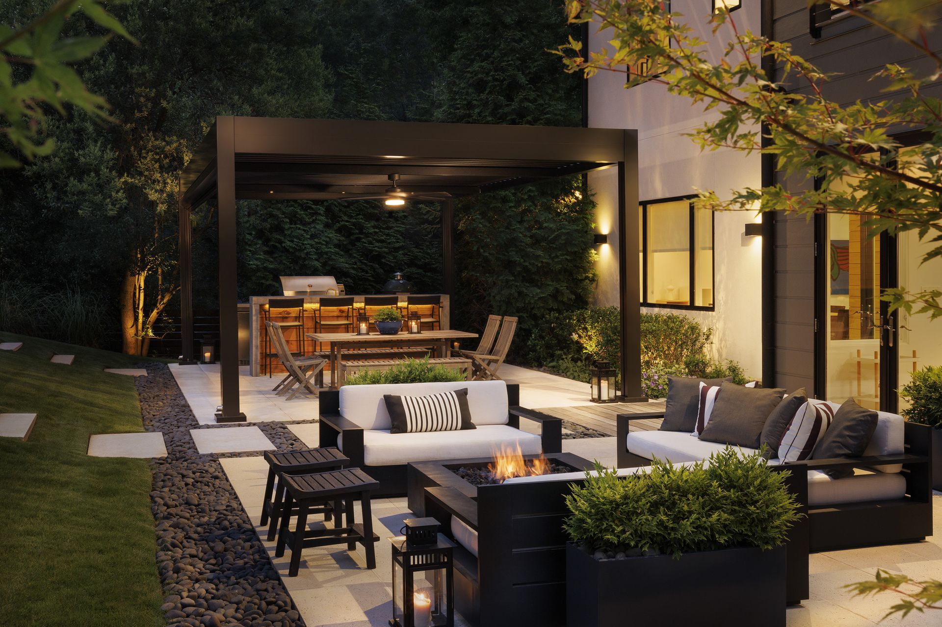 Outdoor patio with dining and seating areas; dark wood pergola, sofas, fire pit, and lush greenery.