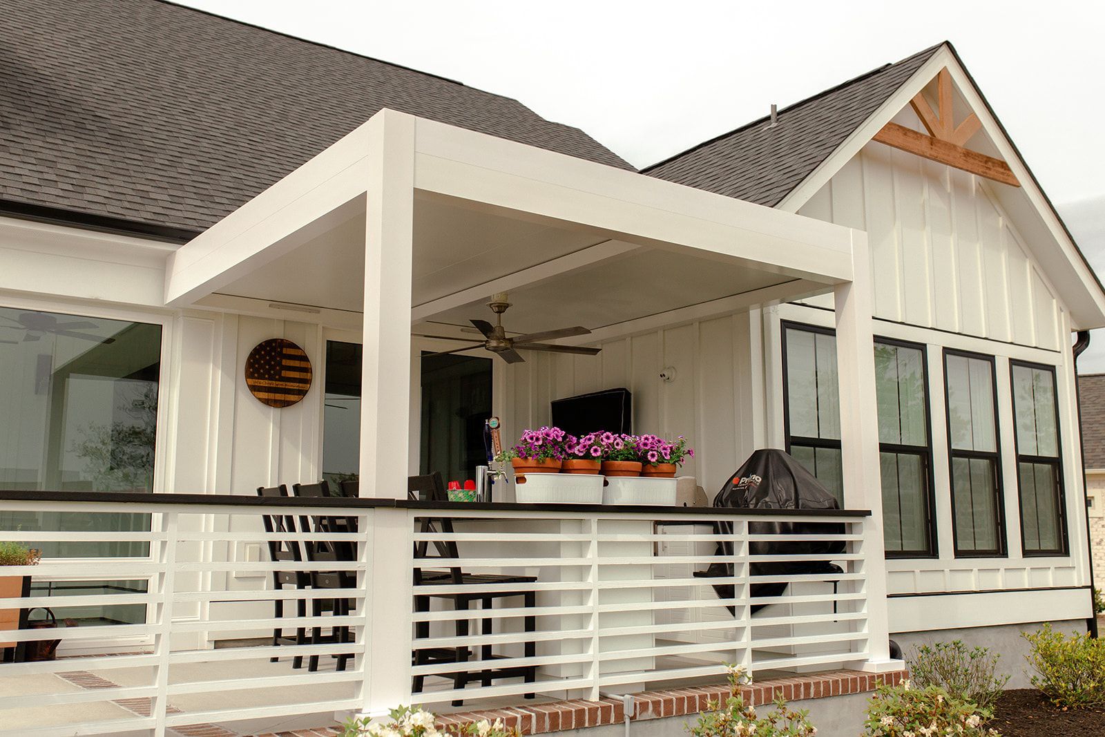 White farmhouse with patio and pergola. White railing, black windows, gray roof, outdoor seating, and potted flowers.