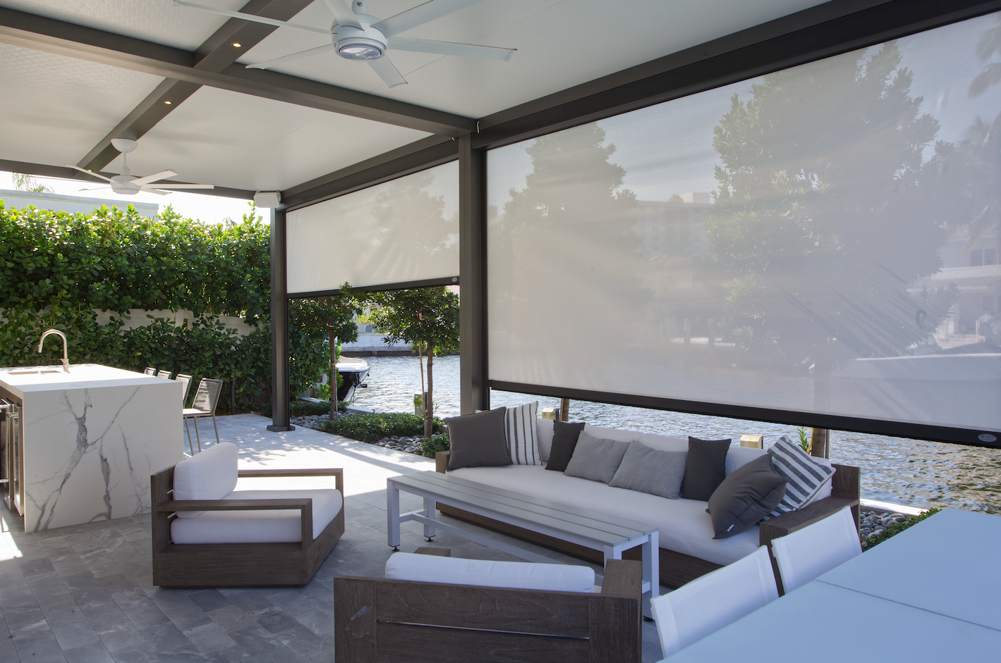 Outdoor patio with white sun shades, seating, and marble-top kitchen overlooking a body of water.