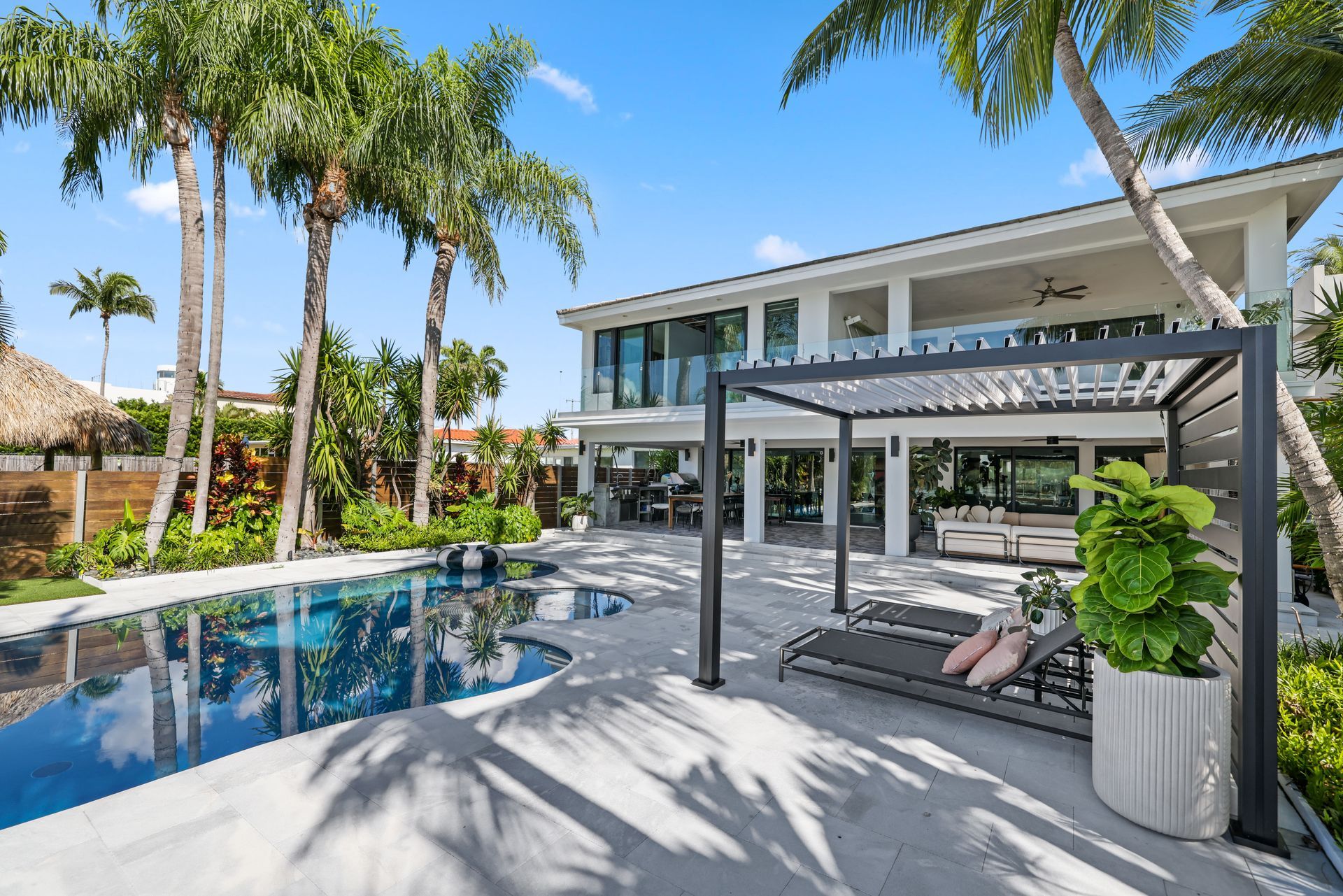 Backyard with pool, modern home, palm trees, pergola with lounge chairs on a sunny day.