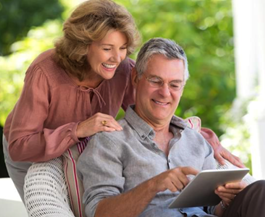 Older couple smiles while looking at a tablet outdoors. Woman rests hand on man's shoulder.