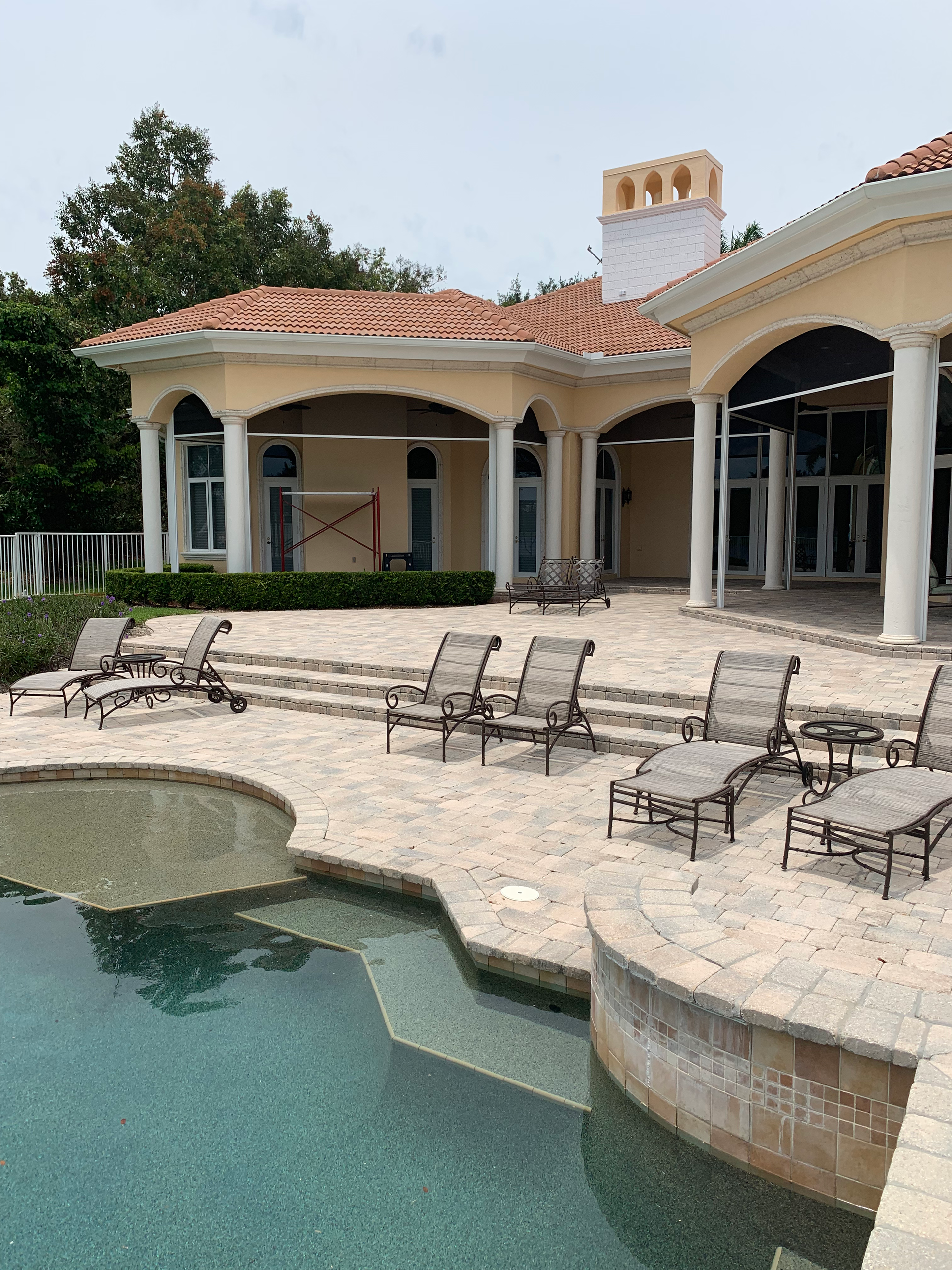 Poolside view of a yellow home with white pillars, a pool, and lounge chairs under an overcast sky.