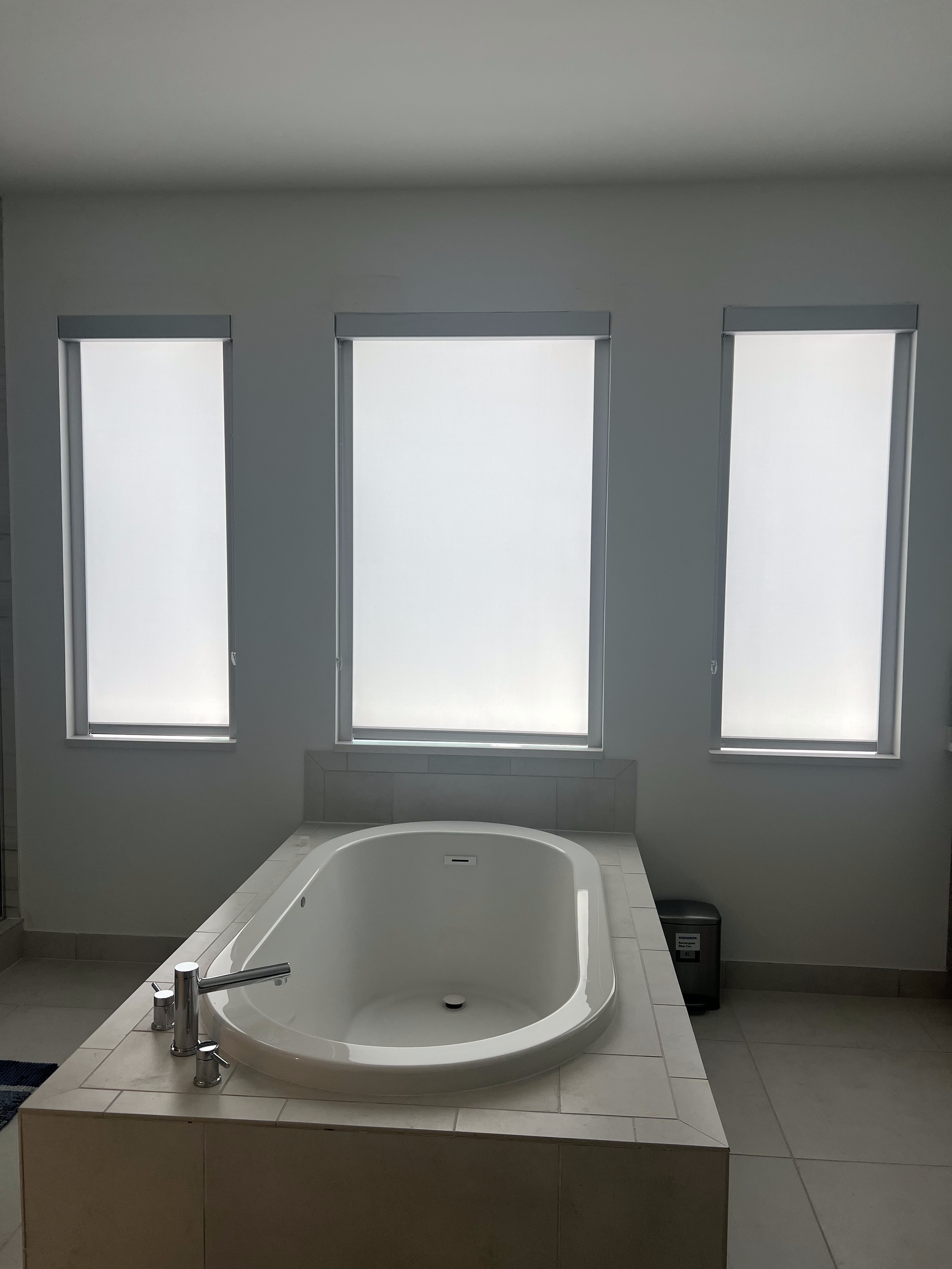 Bathroom with a white bathtub, three windows with blinds, and tiled flooring.