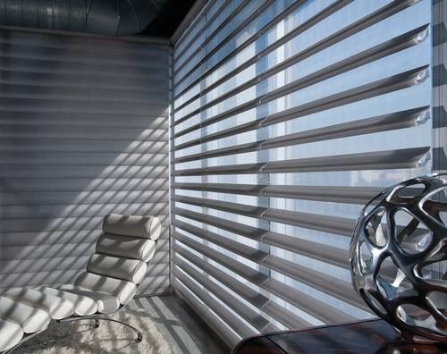 A modern room with layered blinds, a chaise lounge, and a decorative sphere. Sunlight streams in, casting shadows.