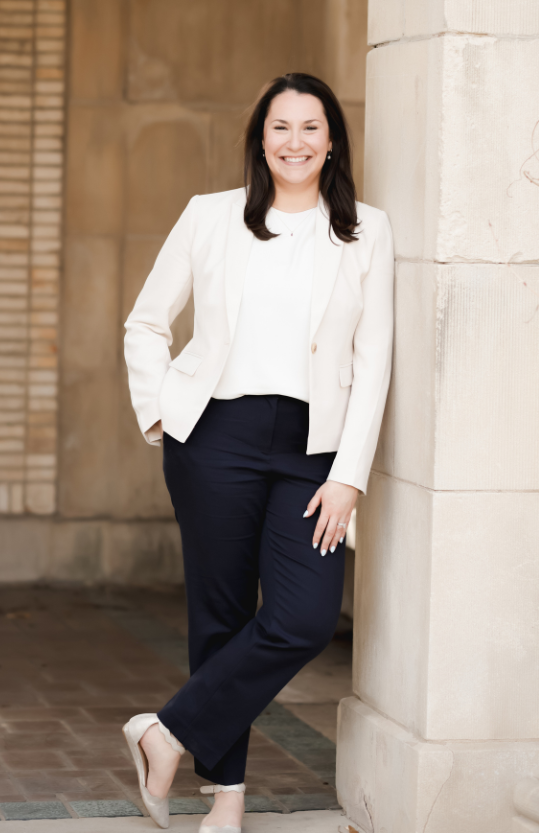 Woman in blazer smiles, sitting on chair against a gray backdrop.