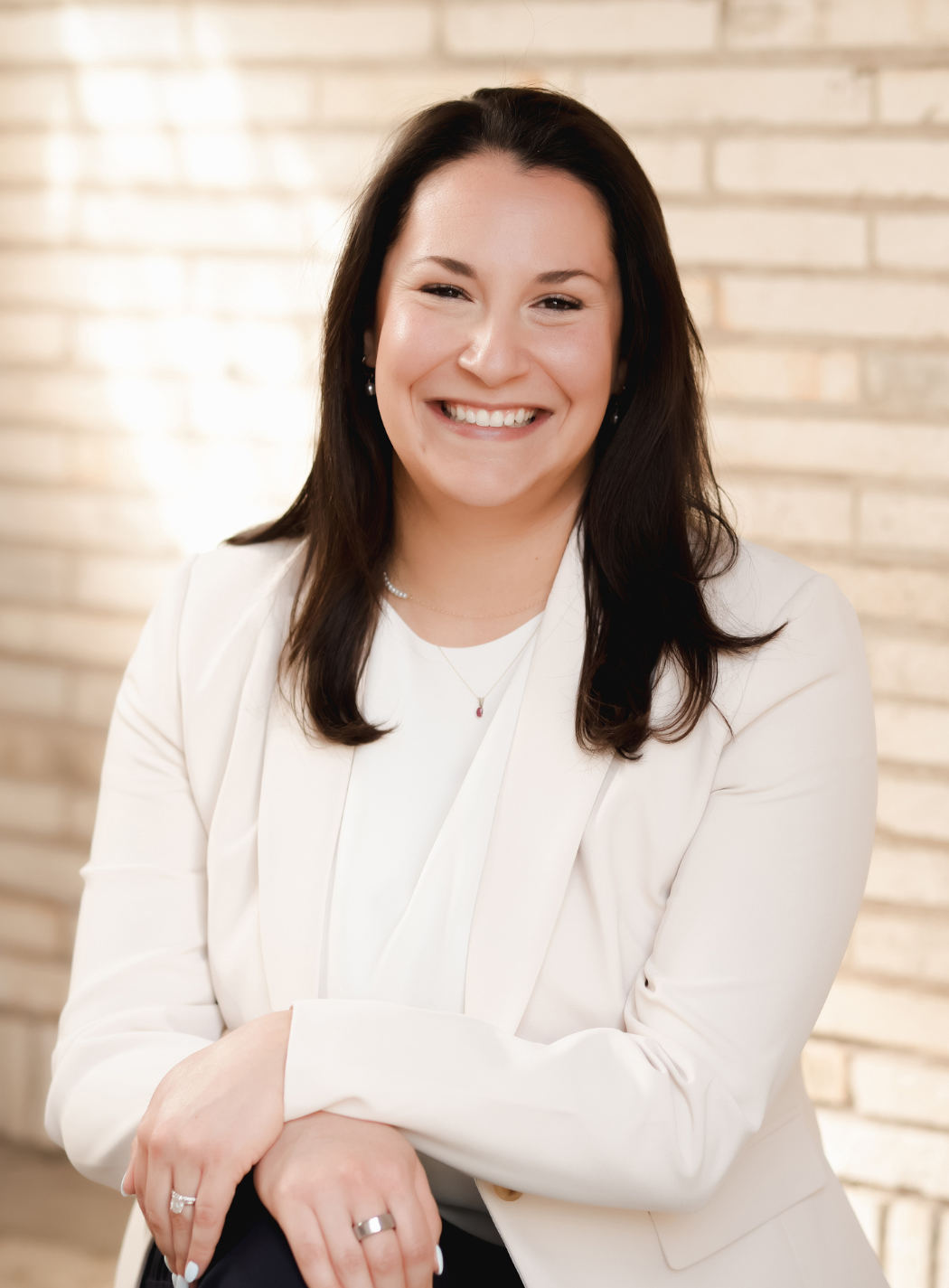 Woman in business attire smiles, sitting in a chair, hand near face, against a grey background.