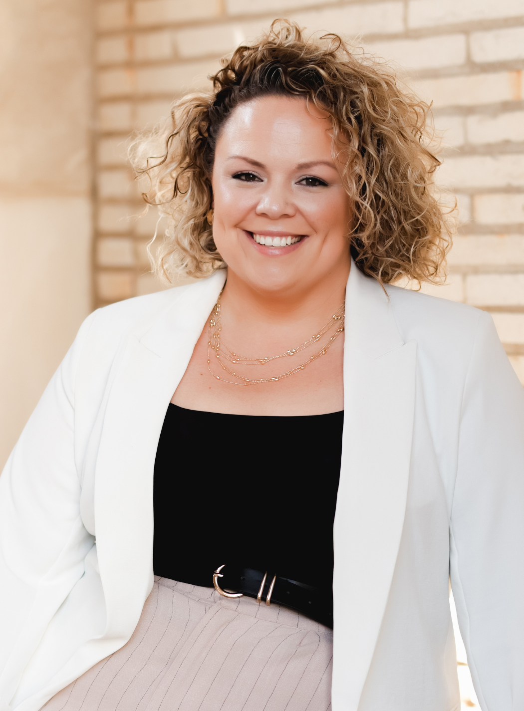 Woman with curly hair smiles, seated, wearing black, on a chair against a gray background.