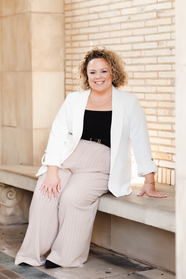 Woman with curly hair, smiling, wearing a black sweater, seated against a grey background.