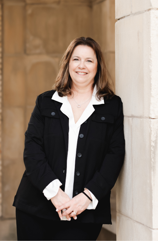 Woman with brown hair, wearing a blue button-down shirt, smiles, leaning on the back of a chair, against a gray wall.