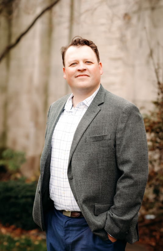 Man in blazer and jeans sits on a chair, looking forward against a gray backdrop.