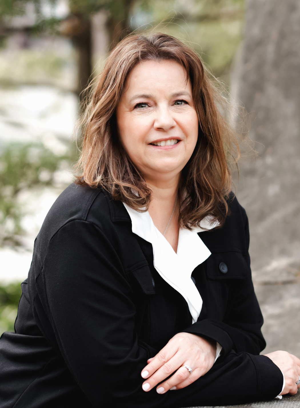 Woman in blue button-down shirt smiles, leaning on the back of a light chair against a gray background.