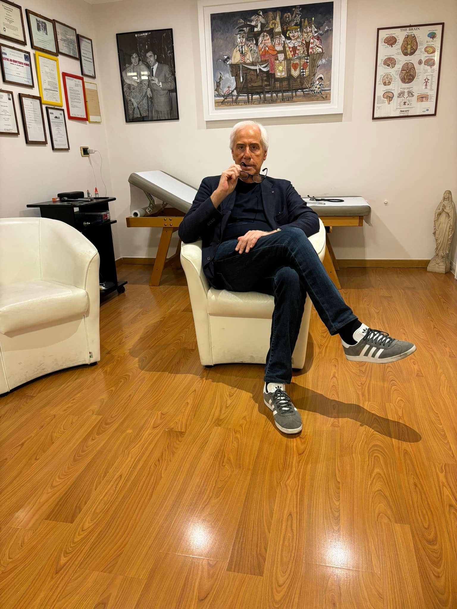 Persona seduta su una sedia bianca in una stanza con quadri incorniciati e un pianoforte, gambe incrociate, mano sul mento.