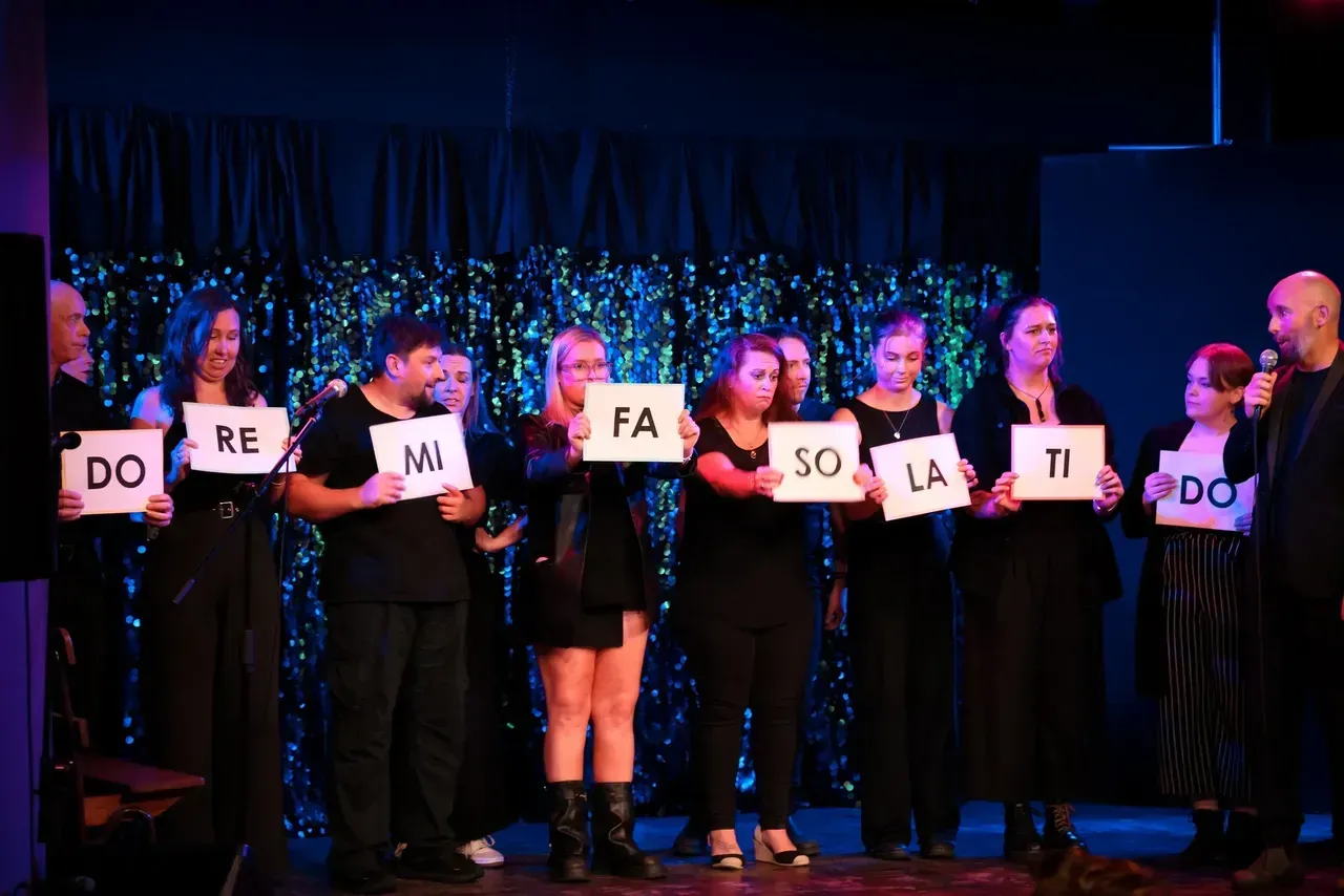People holding white note cards spelling “DO RE MI FA SOL LA TI DO” on a stage with blue lights