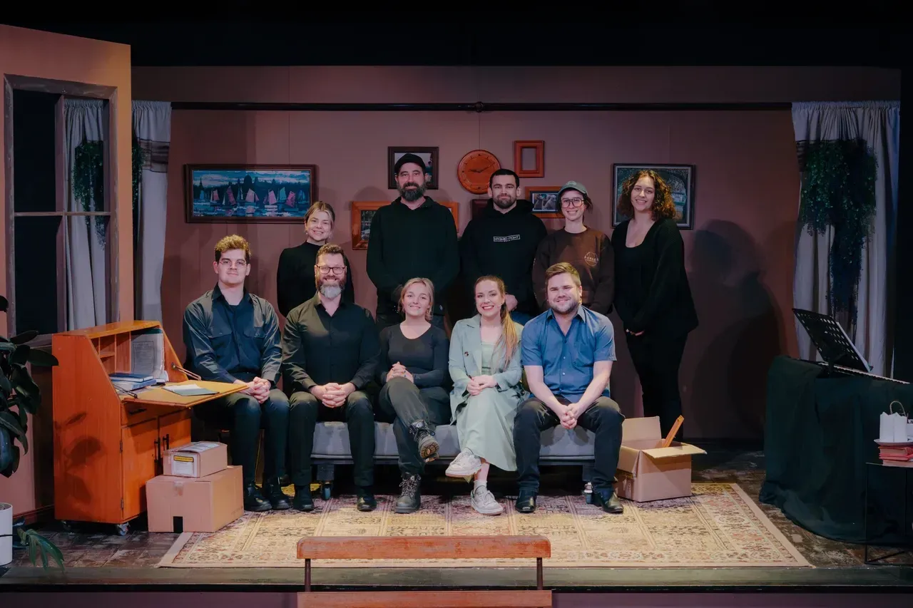 Group posing in a cozy living room set, seated and standing around a couch with warm stage lighting.