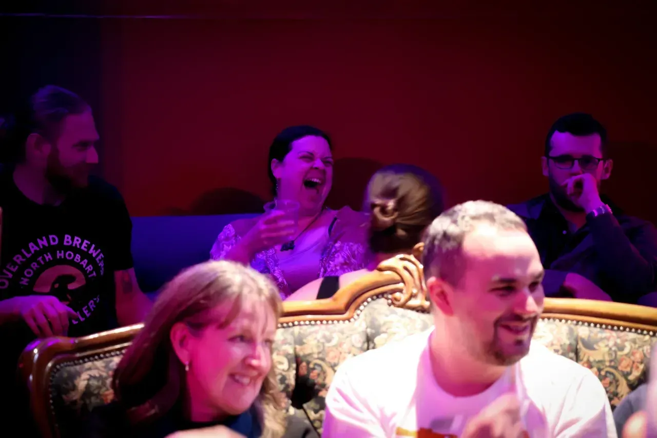 Audience laughing and applauding in a dimly lit venue with purple and red lighting