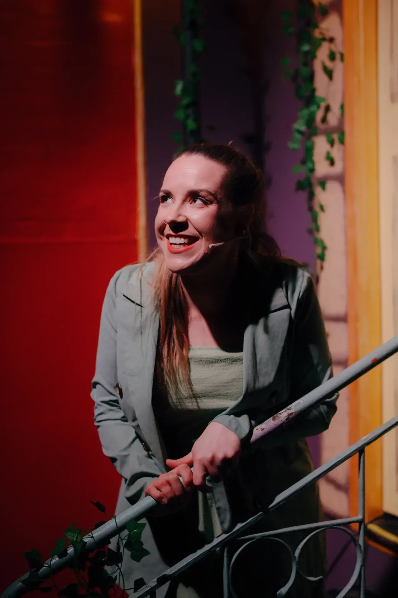 Smiling woman in a gray jacket on a staircase at night, lit by warm red and purple lights.