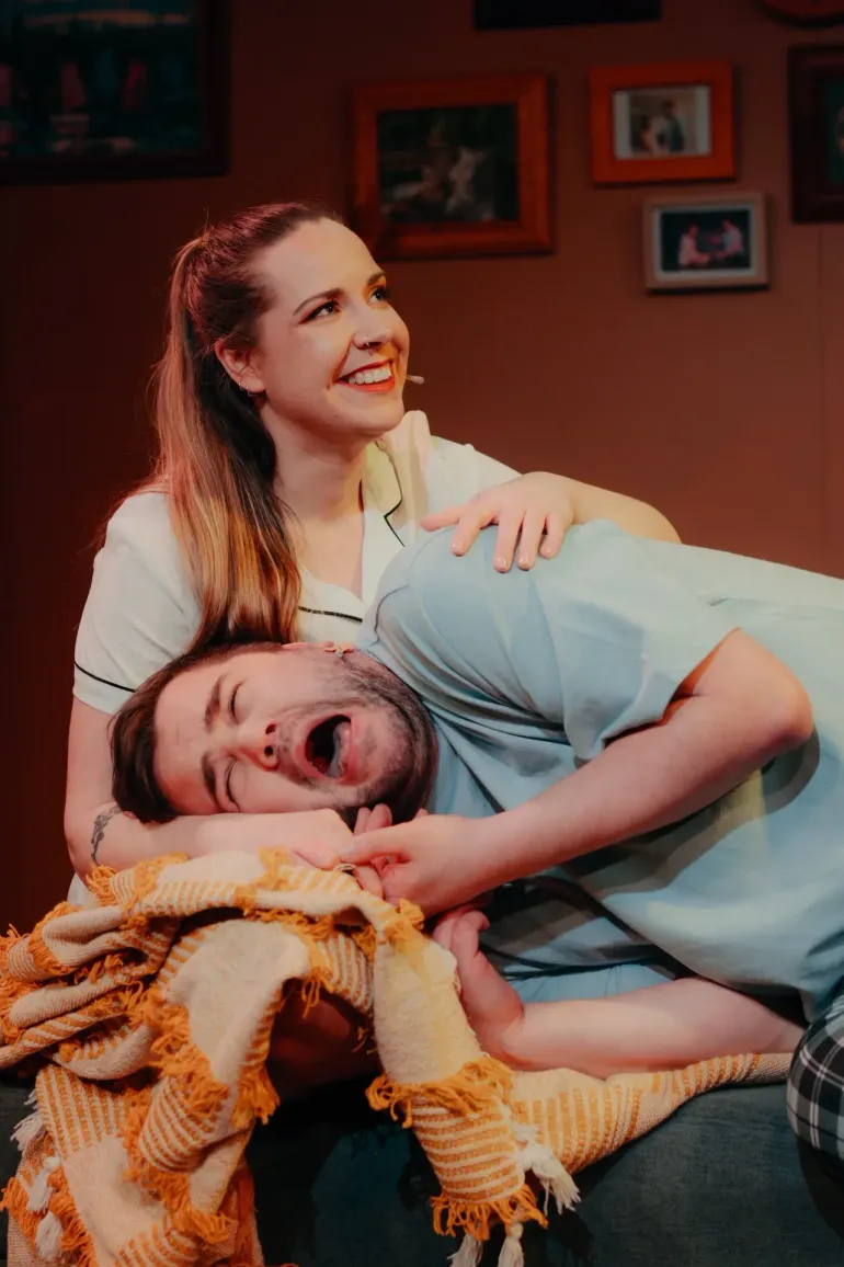 Smiling woman leaning over a man lying on a couch with a blanket, in a warmly lit living room.