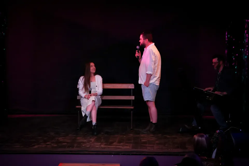 Two people on a dimly lit stage, one seated on a bench and one standing nearby.