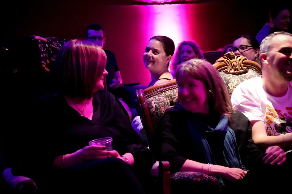 Audience seated in theater, chatting under pink and purple stage lights.