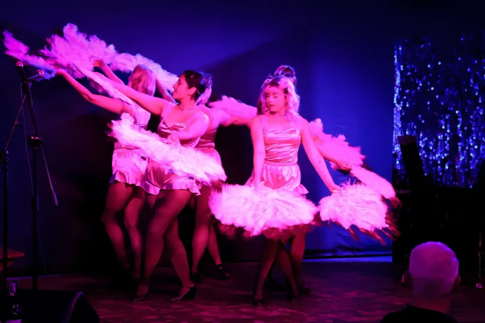 Three dancers in pink feathered costumes performing on a dark stage under purple lighting