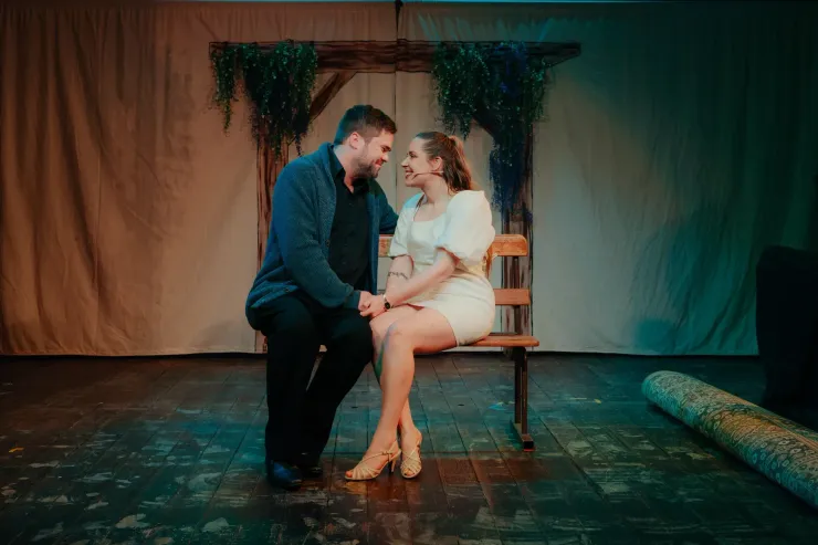 Couple in formal wear sitting close on a chair on a stage, smiling under soft dramatic lighting