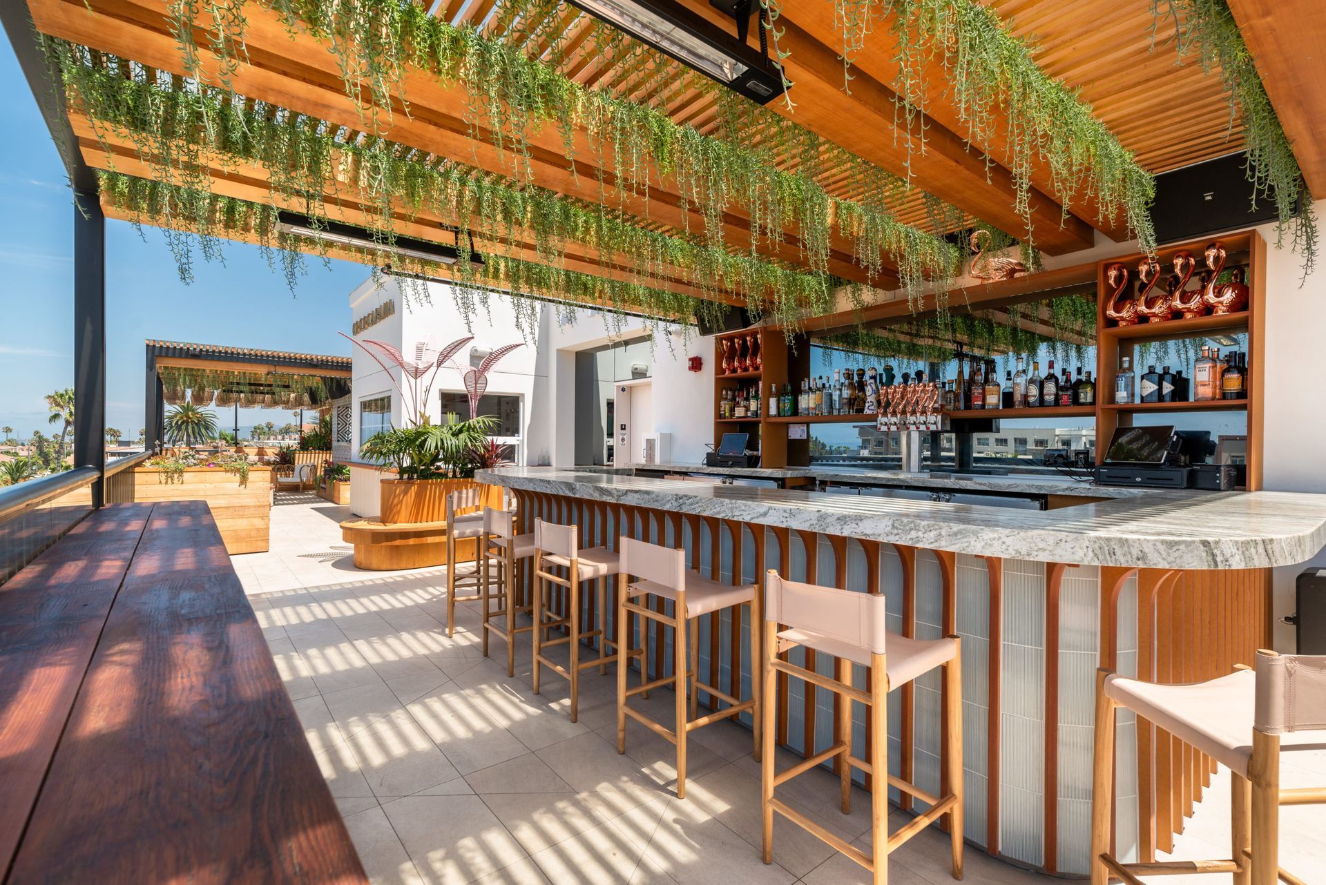 Open-air bar with wooden stools, a marble counter, and hanging greenery under a slatted pergola