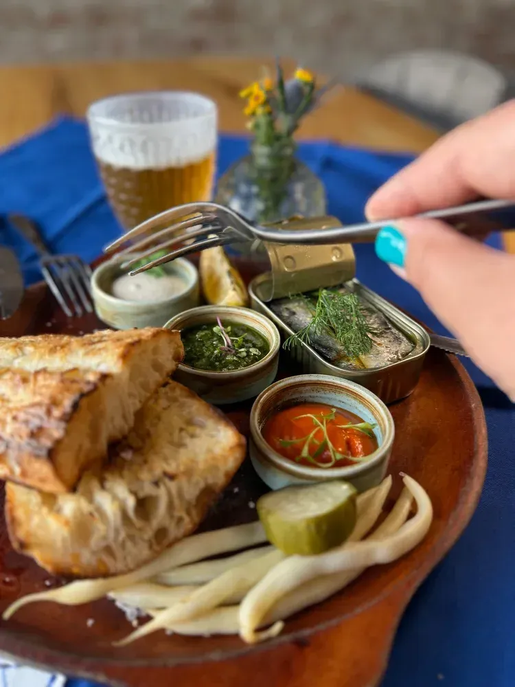 Hand grabbing a wooden platter with bread, pickles, dips, and a beer on a table